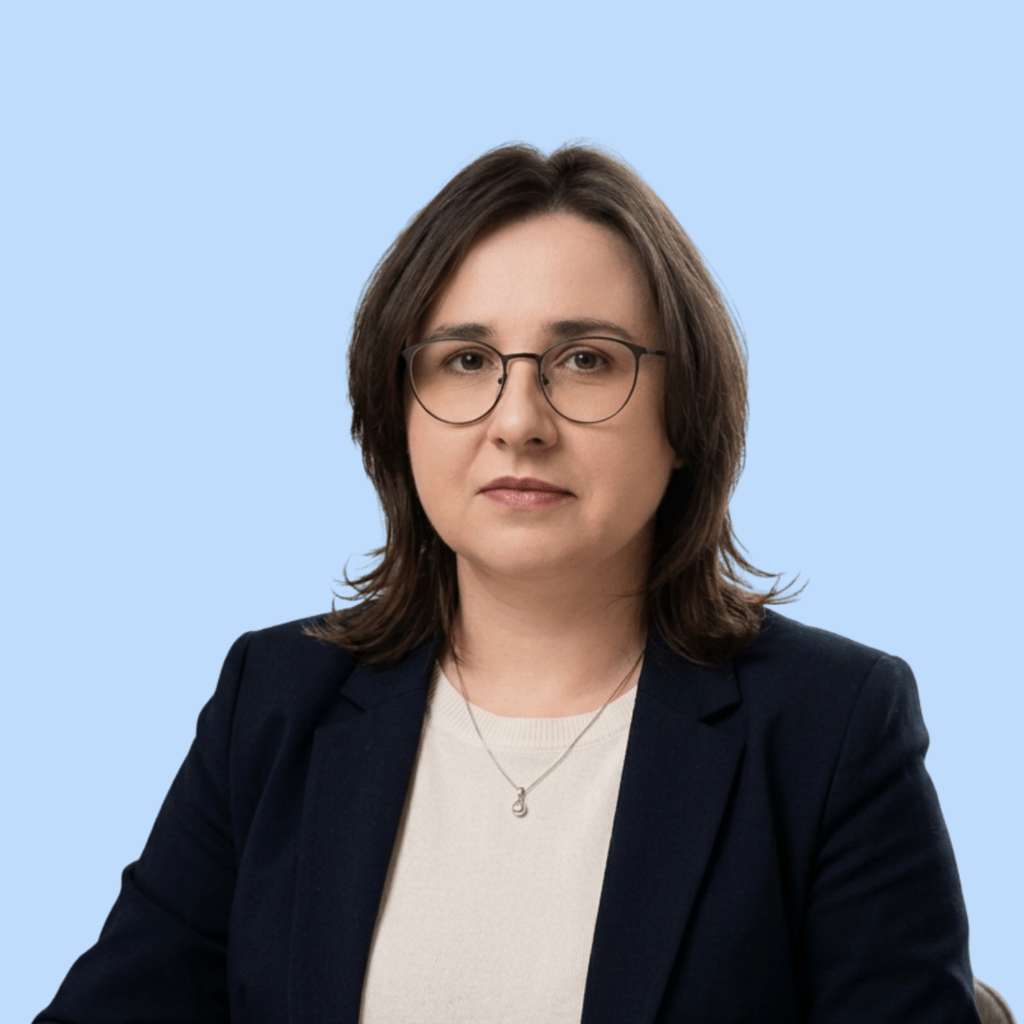 lekarz psychiatra aleksandra mazurek. Zdjęcie przedstawia lekarkę psychiatrę w wieku około 40 lat, w okularach o ciemnych oprawkach i z ciemnymi włosami do ramion, delikatnie uśmiechającą się do kadru. Kobieta ubrana jest w elegancki outfit w stylu smart casual – granatową marynarkę i jasny sweter.